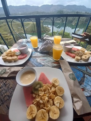 Mesa com itens de café da manhã, incluindo frutas, cereais e bebidas, com paisagem de montanhas ao fundo.