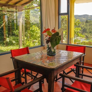 A dining area with a mosaic table, red chairs, and a view of greenery through large windows. - Casa Encuentro Ecolodge (Guatapé)