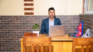A person seated at a wooden desk with a laptop, a small potted plant, and a flag on the wall behind. - Hotel Hidden Kingdom & Spa (Pokhara)