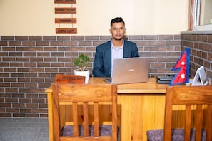 Eine Person sitzt an einem Holztisch, auf dem ein Laptop steht. Außerdem sind ein kleiner Blumentopf und eine Flagge an der Wand im Hintergrund zu sehen.