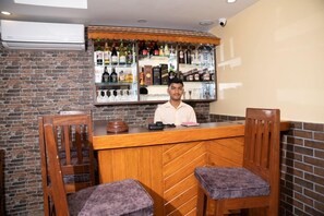 Zona de bar con una barra de madera, dos taburetes acolchados y una variedad de botellas y vasos en estanterías detrás de la barra.