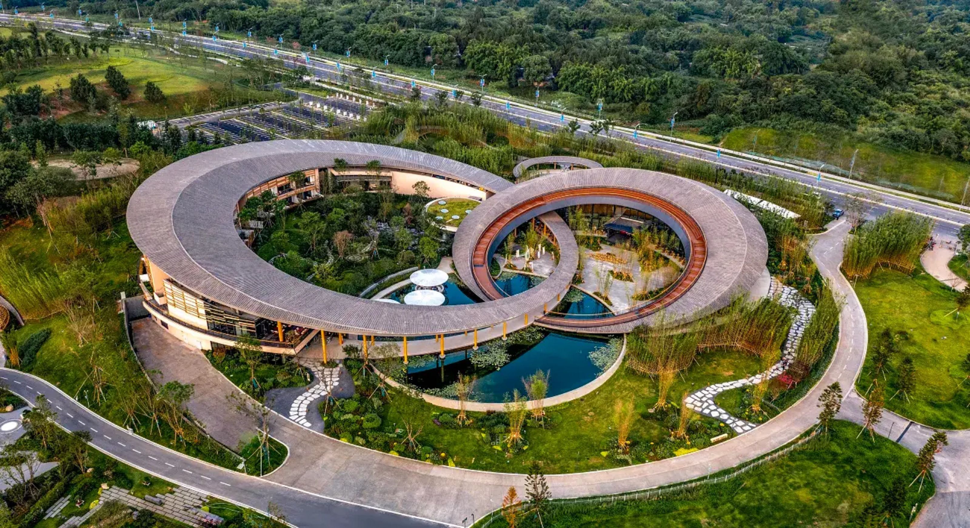 Een rond gebouw met een centraal gelegen zwembad, omgeven door aangelegde tuinen en een bochtige weg.