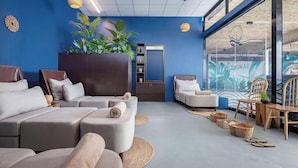 A modern spa room with a blue wall, a large plant, and a glass wall with a tropical mural. There are multiple seating areas with cushions and a wooden cabinet. - Summer Bay Beach Club and Cabins Sihanoukville by Preference (Prey Nob)