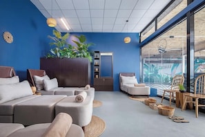 Un spa moderno con pared azul, una planta grande y un ventanal con un mural tropical. Hay varias zonas de estar con cojines y un mueble de madera.