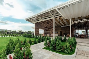 Un edificio moderno con un espacio de asientos techado al aire libre, rodeado de abundante vegetación y un cielo despejado.