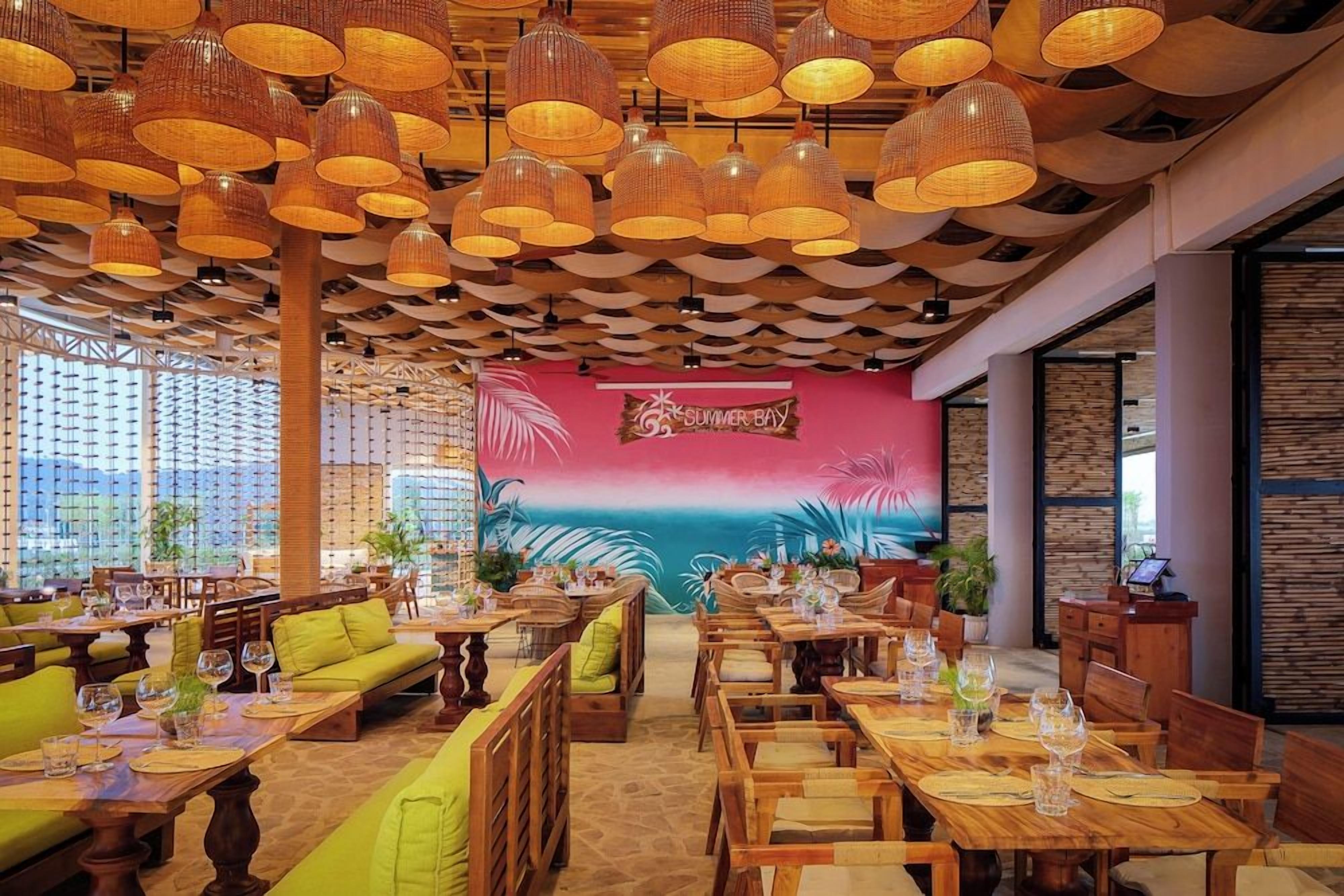 Een restaurant met houten tafels en stoelen, een muurschildering en hangende rieten lampen.
