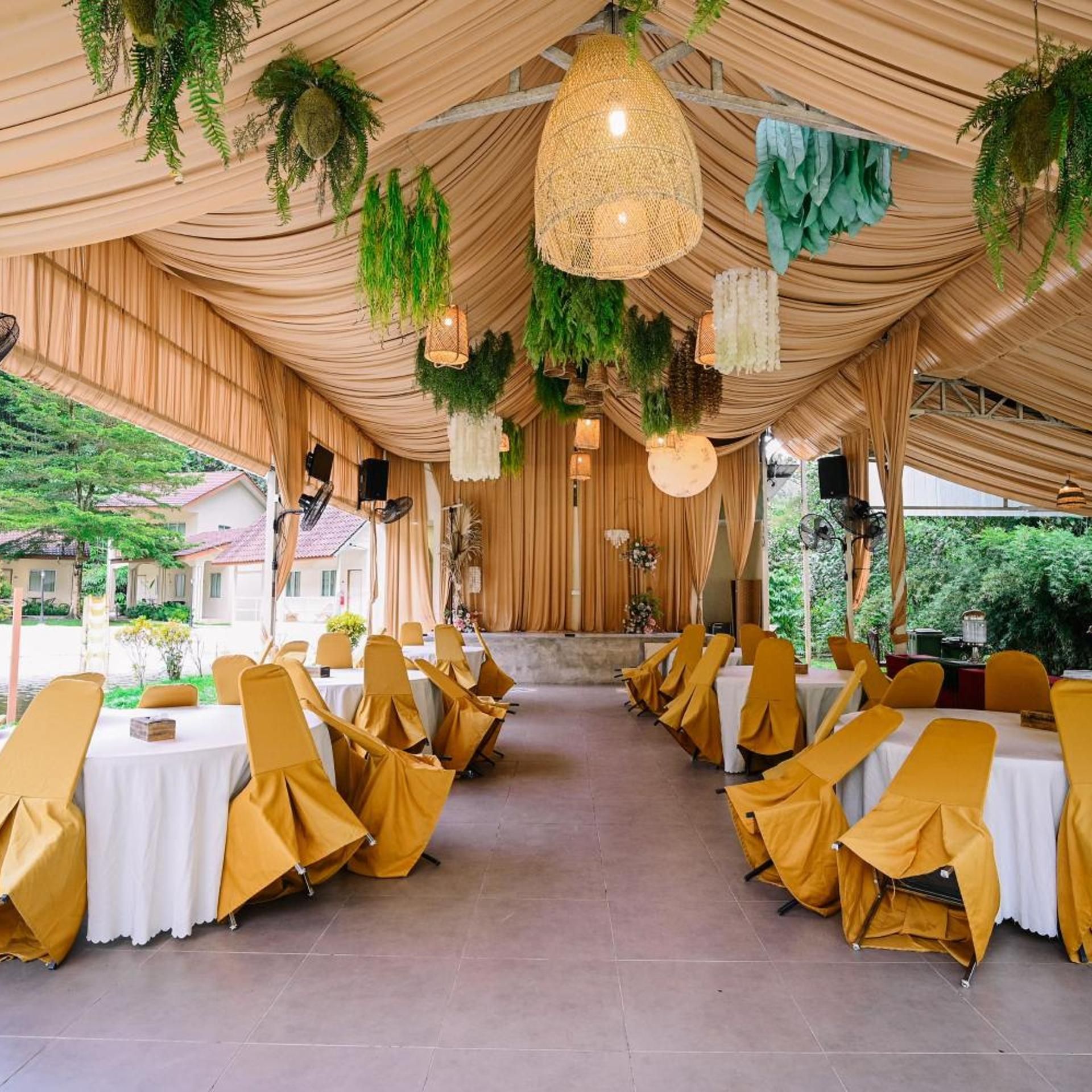 An indoor event space with tables and chairs, draped with golden fabric, and decorated with hanging plants and lights.