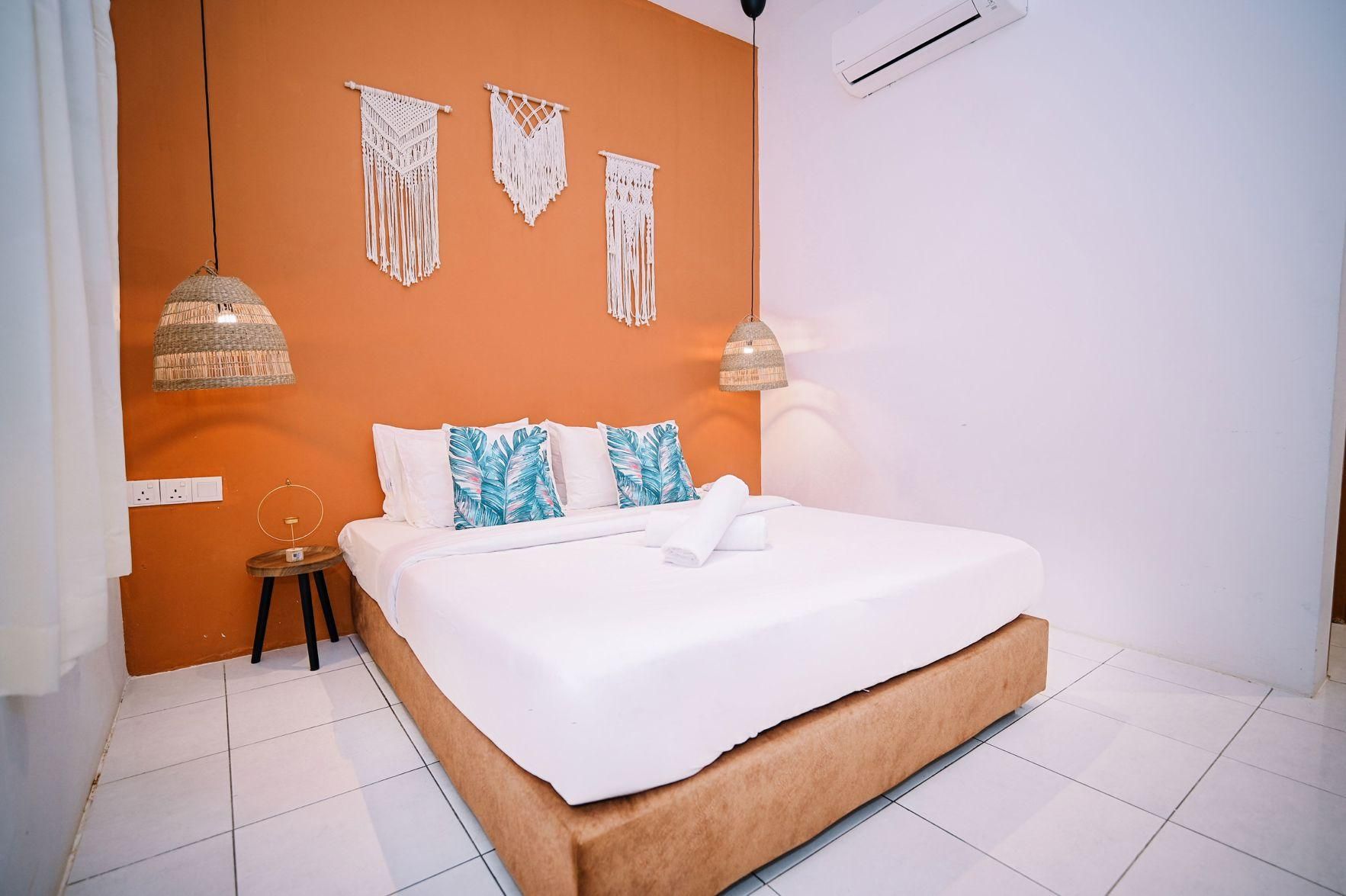 A bedroom with a bed, white bedding, a small bedside table, a wall-mounted air conditioner, and macrame wall hangings.