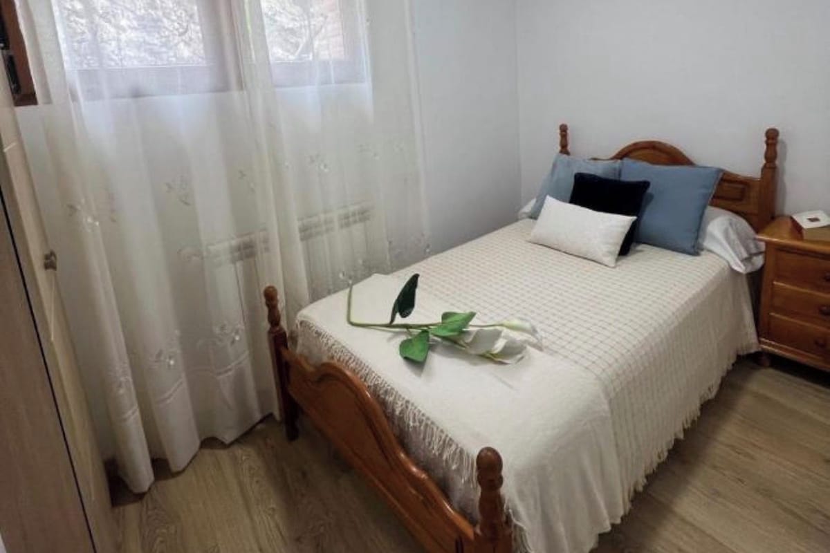 A bedroom with a wooden bed, white bedding, a bedside table, and a window with sheer curtains.