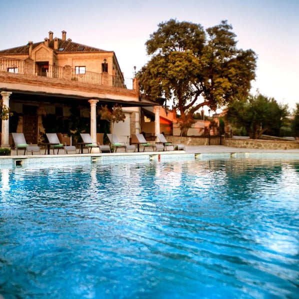A swimming pool with lounge chairs, a building with columns, and a large tree. - Hotel Cigarral Santa Maria (Toledo)