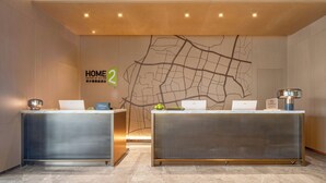 Una recepción de hotel con dos recepcionistas, un mapa en la pared y un cartel que indica 'HOME 2' y 'Hilton Honors'.