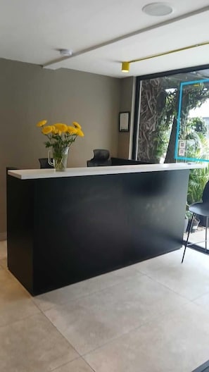 Un mostrador de recepción con un florero de flores amarillas, una silla negra y vistas al exterior.