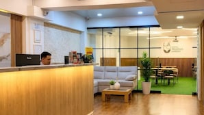 A hotel reception area with a wooden reception desk, a seating area with a sofa and coffee table, and a dining area visible through glass doors. - Inactivo Hotel Natura Express (Medellín)