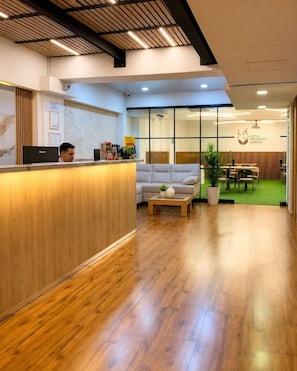 A hotel reception area with a wooden reception desk, a seating area with a sofa and coffee table, and a dining area visible through glass doors. - Inactivo Hotel Natura Express (Medellín)