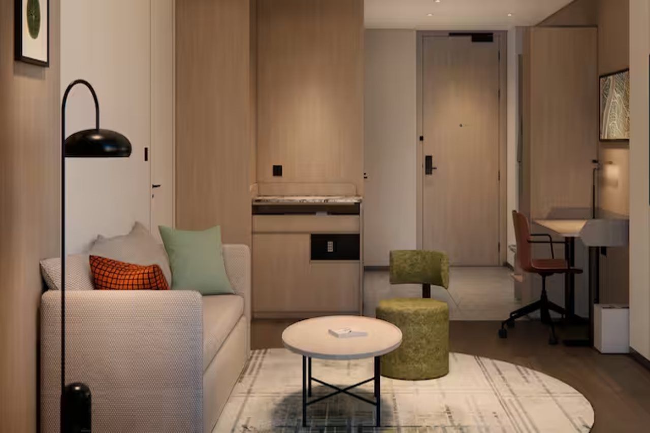 A modern living room with a sofa, a round coffee table, and a green ottoman. There is a kitchen area with cabinets and a countertop visible in the background.