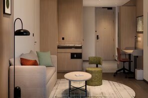 A modern living room with a sofa, a round coffee table, and a green ottoman. There is a kitchen area with cabinets and a countertop visible in the background. - Hilton Garden Inn Tianjin Jinnan (Tianjin)