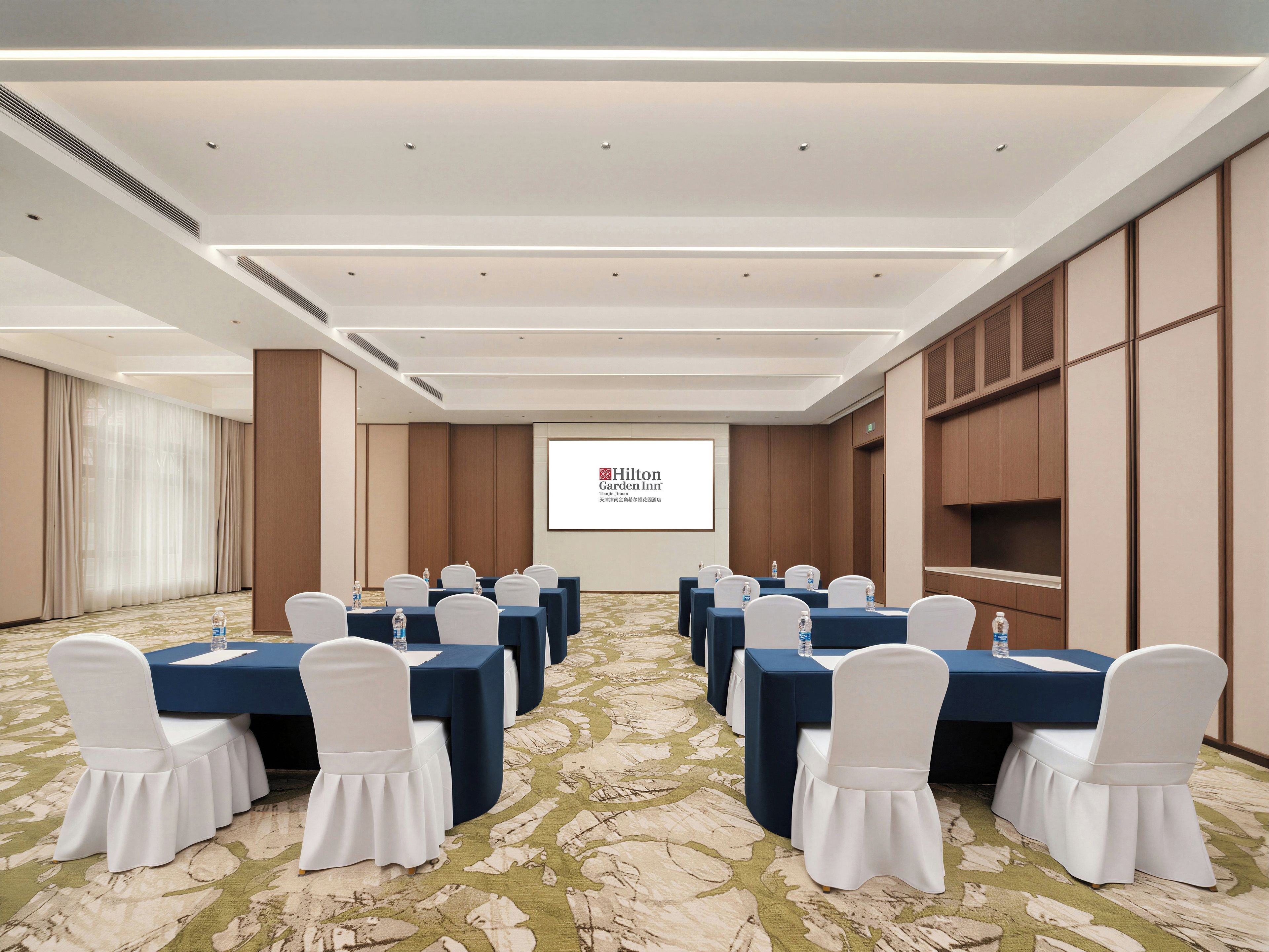 Sala de conferência com mesas, cadeiras e tela de projeção exibindo a marca Hilton Garden Inn.