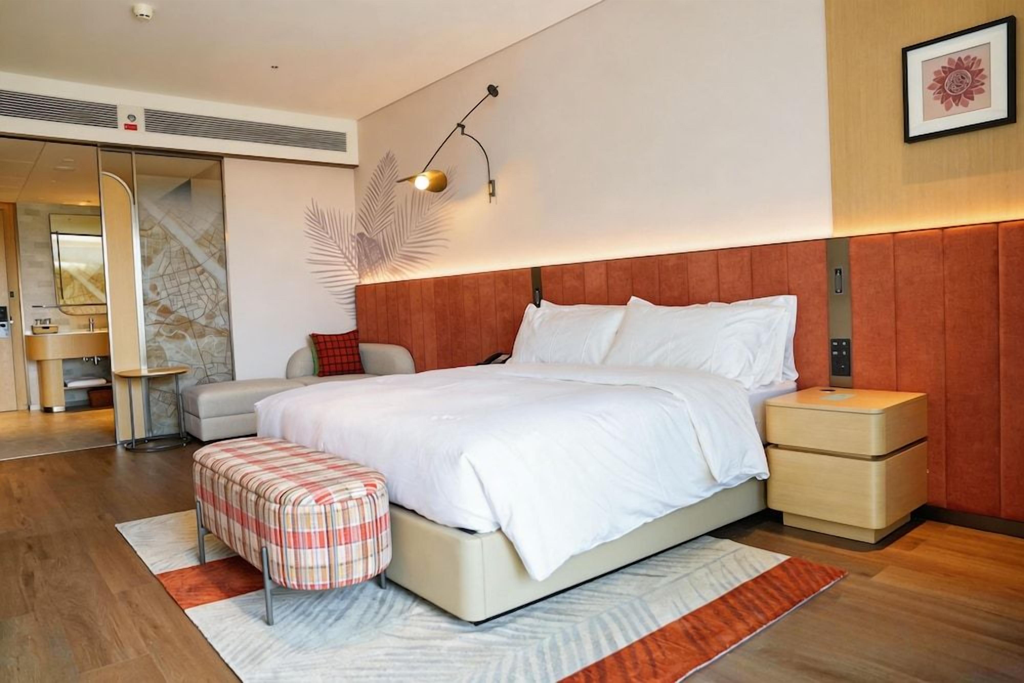 A modern hotel room with a large bed, a bedside table, a nightstand, a bench, and a wall-mounted lamp.