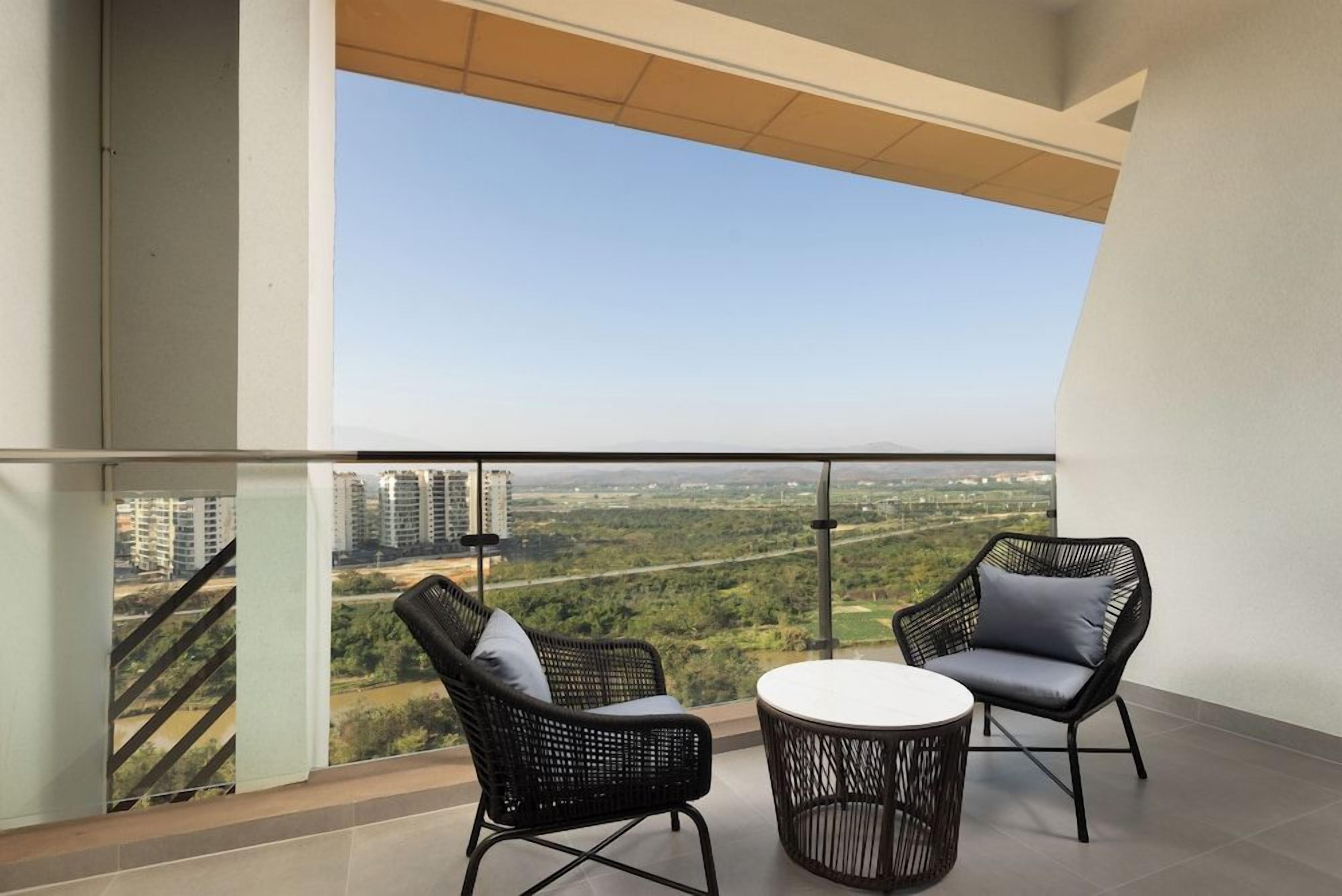 A balcony with two wicker chairs and a small round table, overlooking a cityscape with buildings and greenery.