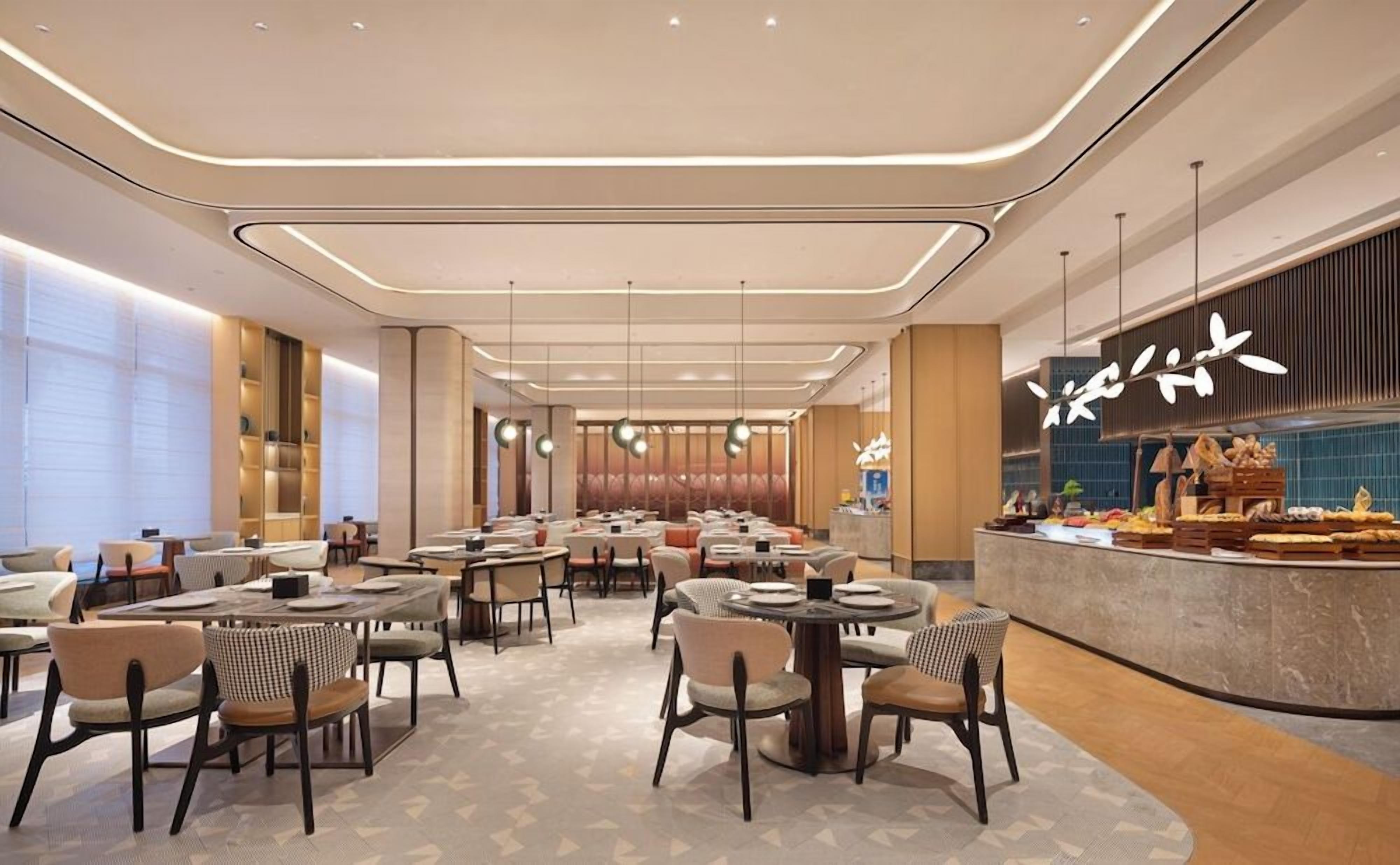 A modern hotel dining area with multiple tables and chairs, a central bar area, and a patterned floor.