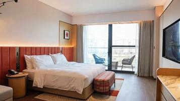 Habitación de hotel con cama, mesita de noche, una pequeña mesa redonda, un banco, televisor y balcón con una silla.