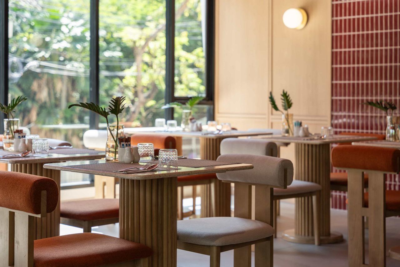 A restaurant with multiple tables and chairs, some with cushions. There are plants on the tables, and the setting appears to be indoors with natural light coming through large windows.