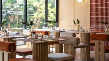 A restaurant with multiple tables and chairs, some with cushions. There are plants on the tables, and the setting appears to be indoors with natural light coming through large windows.
