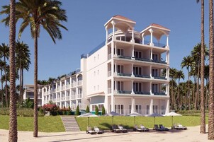 Un hotel de varios pisos con balcones, una zona de piscina con tumbonas y palmeras.