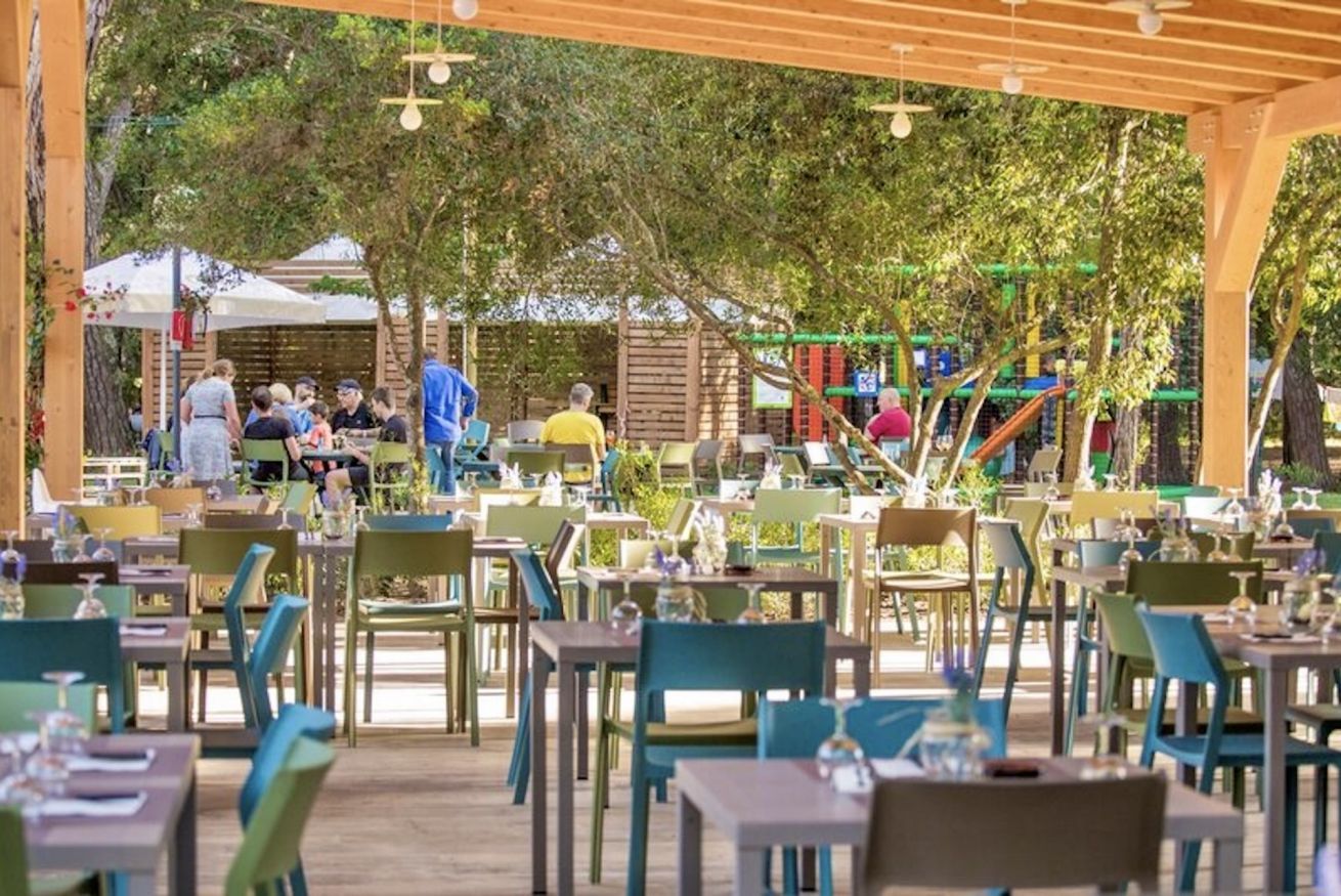 An outdoor dining area with multiple tables and chairs, surrounded by trees and a playground in the background.
