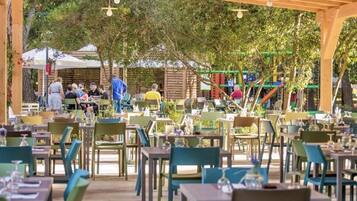 An outdoor dining area with multiple tables and chairs, surrounded by trees and a playground in the background.
