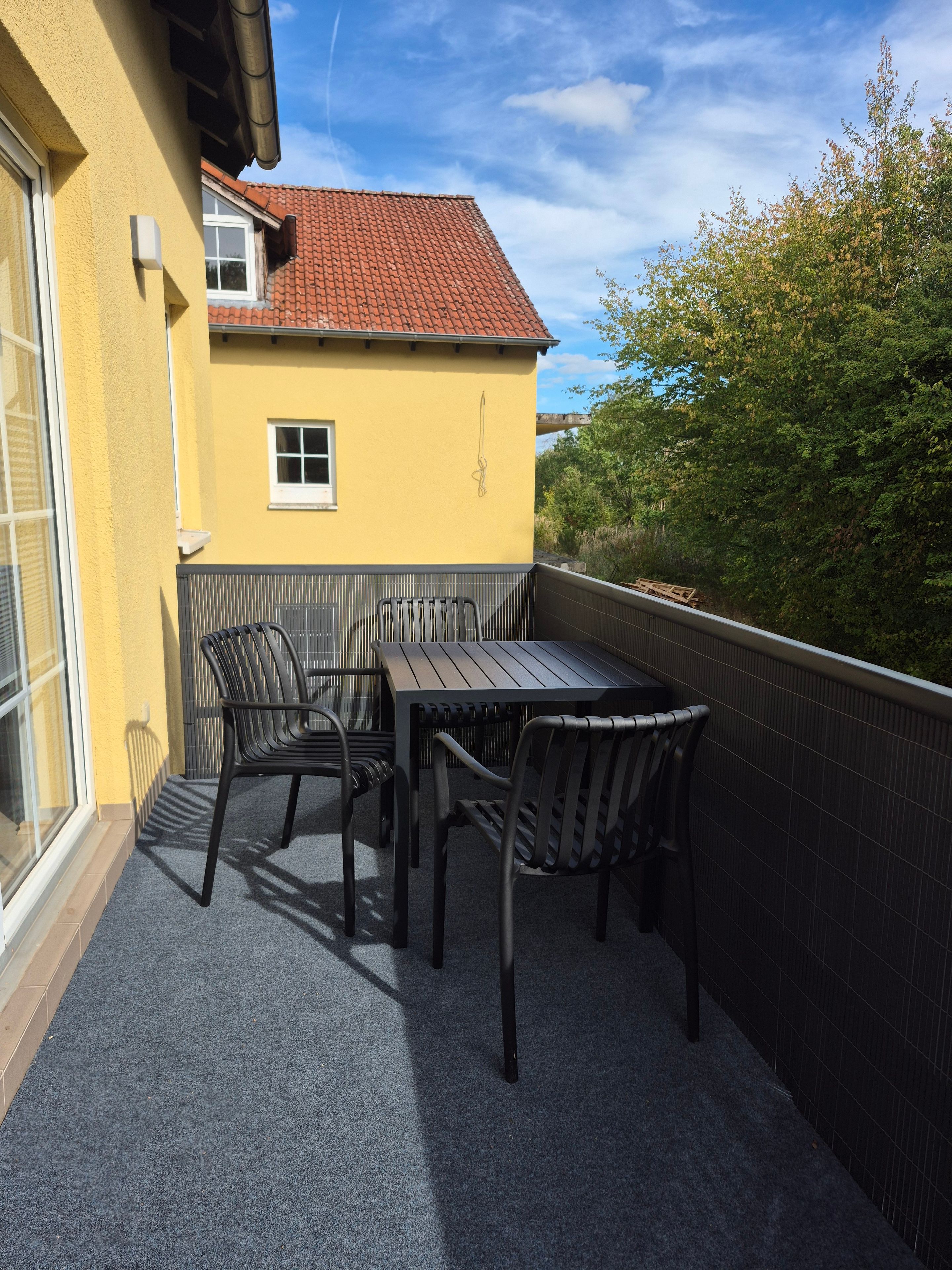 Un balcon avec une table et des chaises, un immeuble jaune avec un toit rouge et une vue sur les arbres.