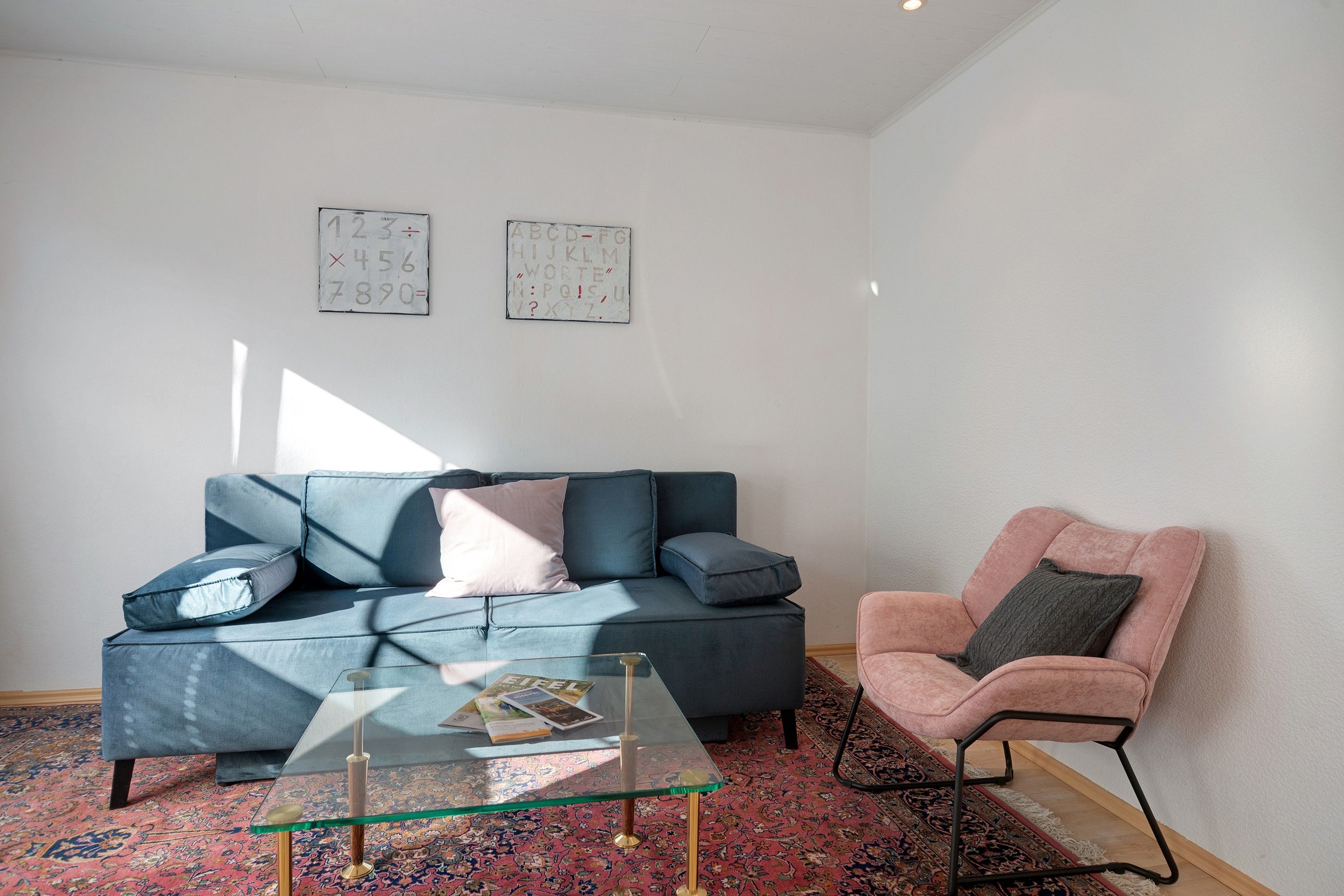 Una sala de estar con un sofá azul, una mesa de centro de vidrio, un sillón rosa y una alfombra estampada.