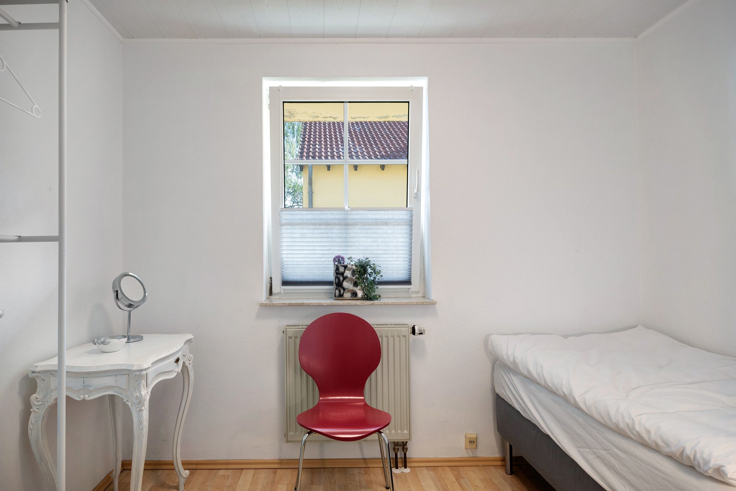 Une petite pièce propre, avec un lit, une chaise, une petite table avec un miroir et une fenêtre donnant sur un immeuble.