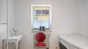 Habitació petita i neta amb llit, cadira, taula petita amb mirall i finestra que dona a un edifici.