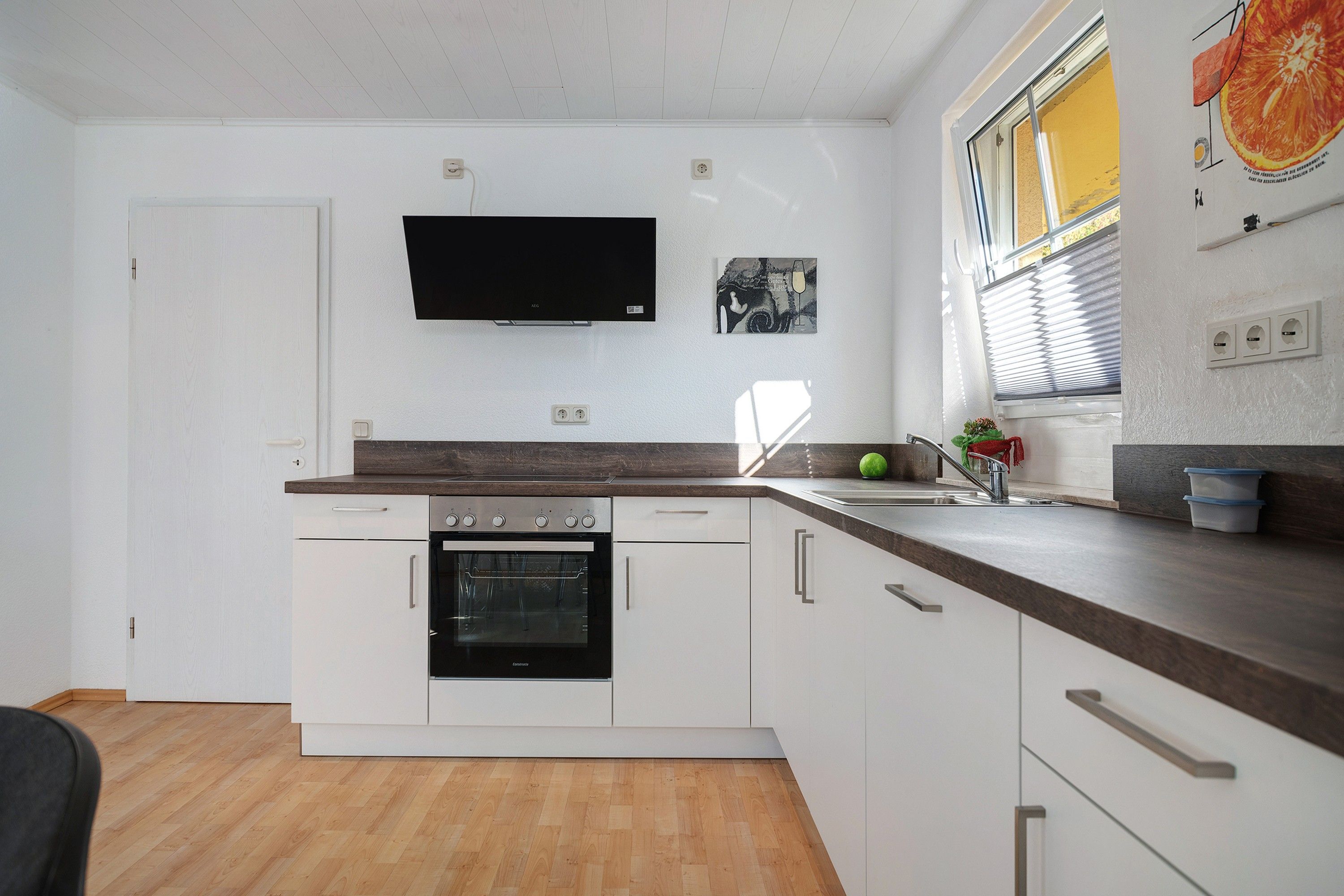 Een moderne keuken met witte kastjes, een ingebouwde oven, een flatscreen televisie en een raam met zonwering.