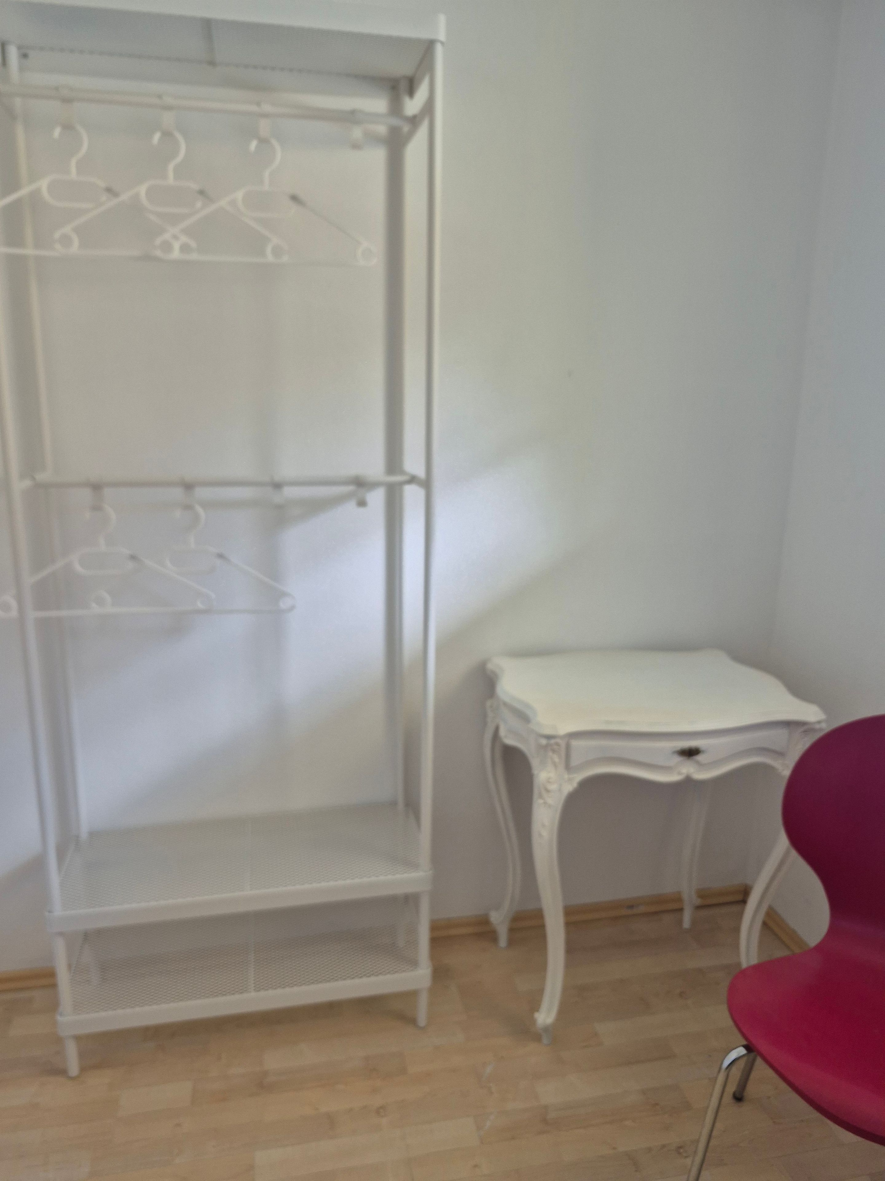 A white metal wardrobe with multiple shelves and hangers, a small white table with a drawer, and a red chair.