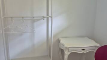 A white metal wardrobe with multiple shelves and hangers, a small white table with a drawer, and a red chair.