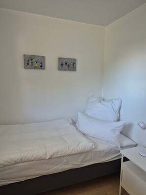 A single-bed room with white bedding, a bedside table with a lamp, and two wall-mounted artwork pieces.