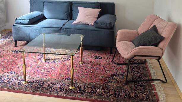 Een woonkamer met een blauwe bank, een glazen salontafel, een roze fauteuil en een kamerkleed met een patroon.