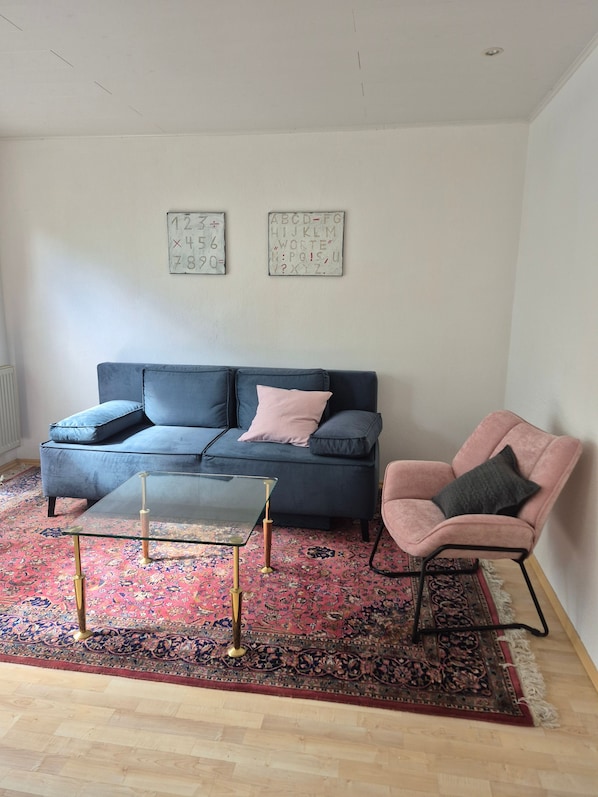 Ein Wohnzimmer mit einem blauen Sofa, einem Glastisch, einem rosa Sessel und einem gemusterten Teppich.