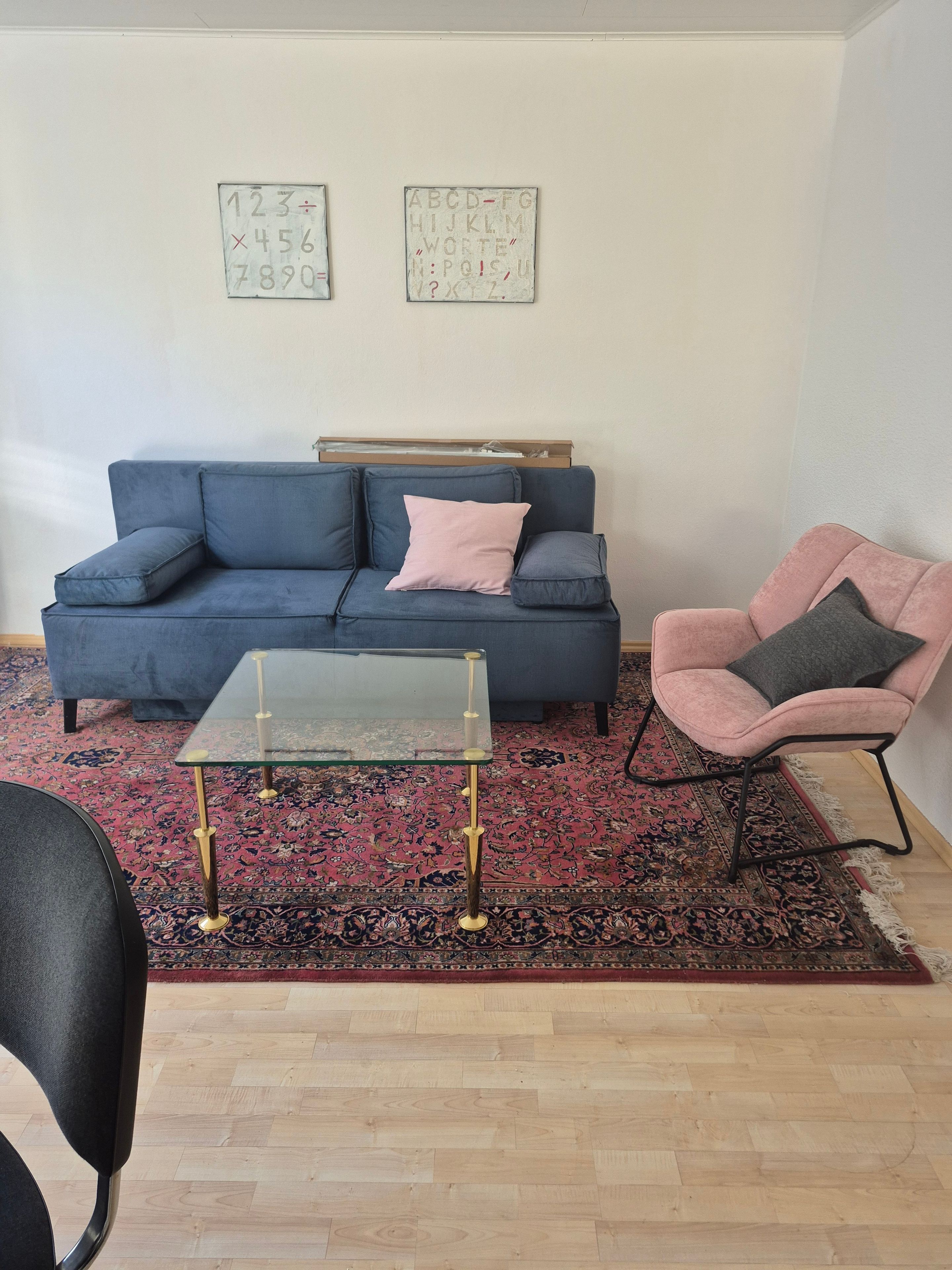 Ein Wohnzimmer mit einem blauen Sofa, einem Glastisch, einem rosa Sessel und einem gemusterten Teppich.