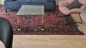 Een woonkamer met een blauwe bank, een glazen salontafel, een roze fauteuil en een vloerkleed met een patroon.