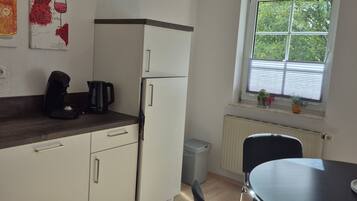 A compact kitchen with white cabinets, a refrigerator, a coffee maker, and a window with blinds.