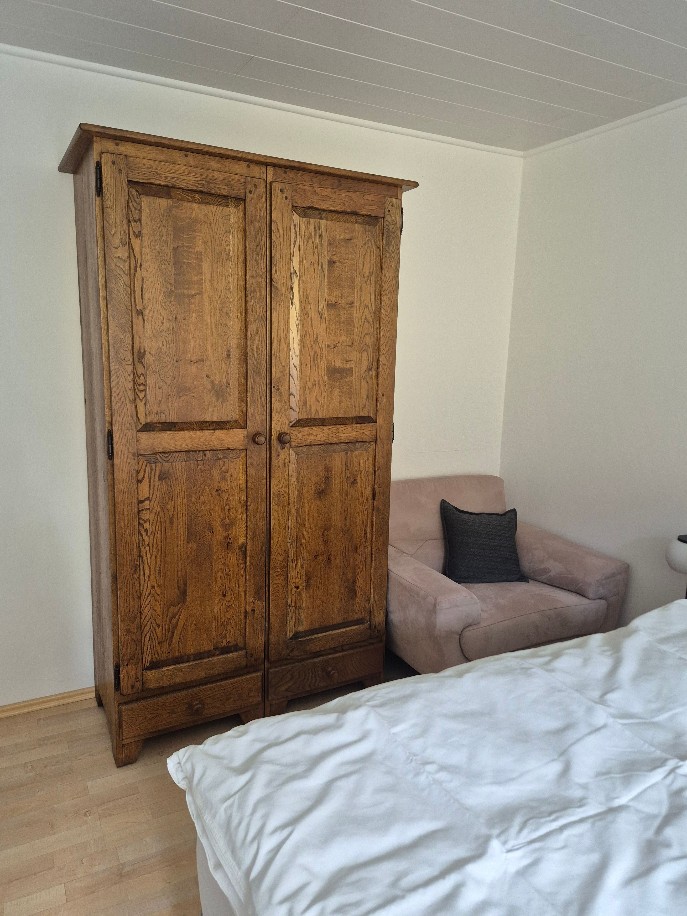 Ein Zimmer mit einem Holzschrank, einem Bett mit weißer Bettwäsche und einem rosafarbenen Sofa mit einem dunkelgrauen Kissen.