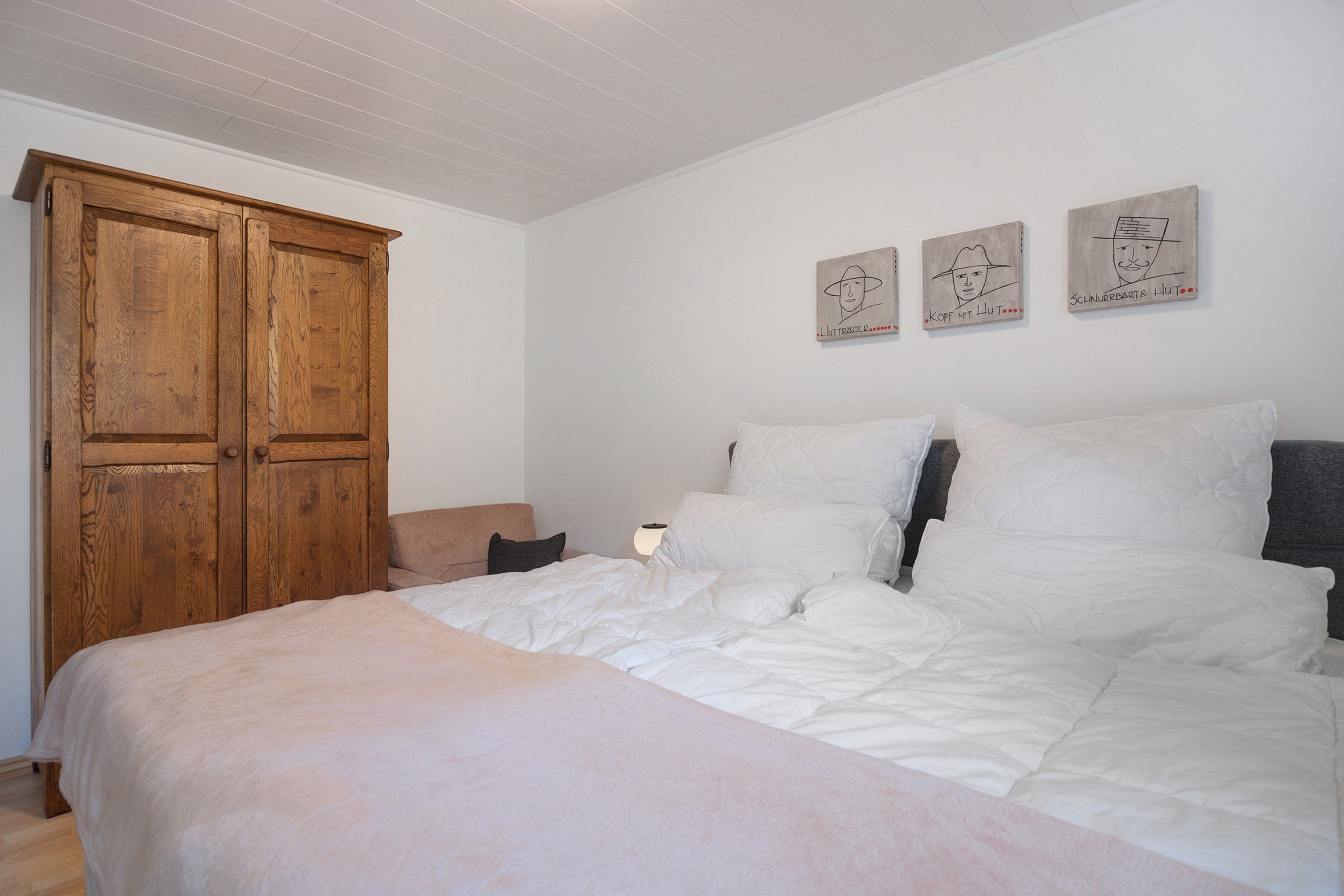 Une chambre avec une grande armoire en bois, un lit avec une literie blanche et trois œuvres d’art accrochées au mur.