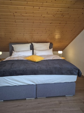 Ein Schlafzimmer mit Schräge, einem Bett mit grauer Bettwäsche und einem gelben Kopfkissen sowie Nachttischlampen.