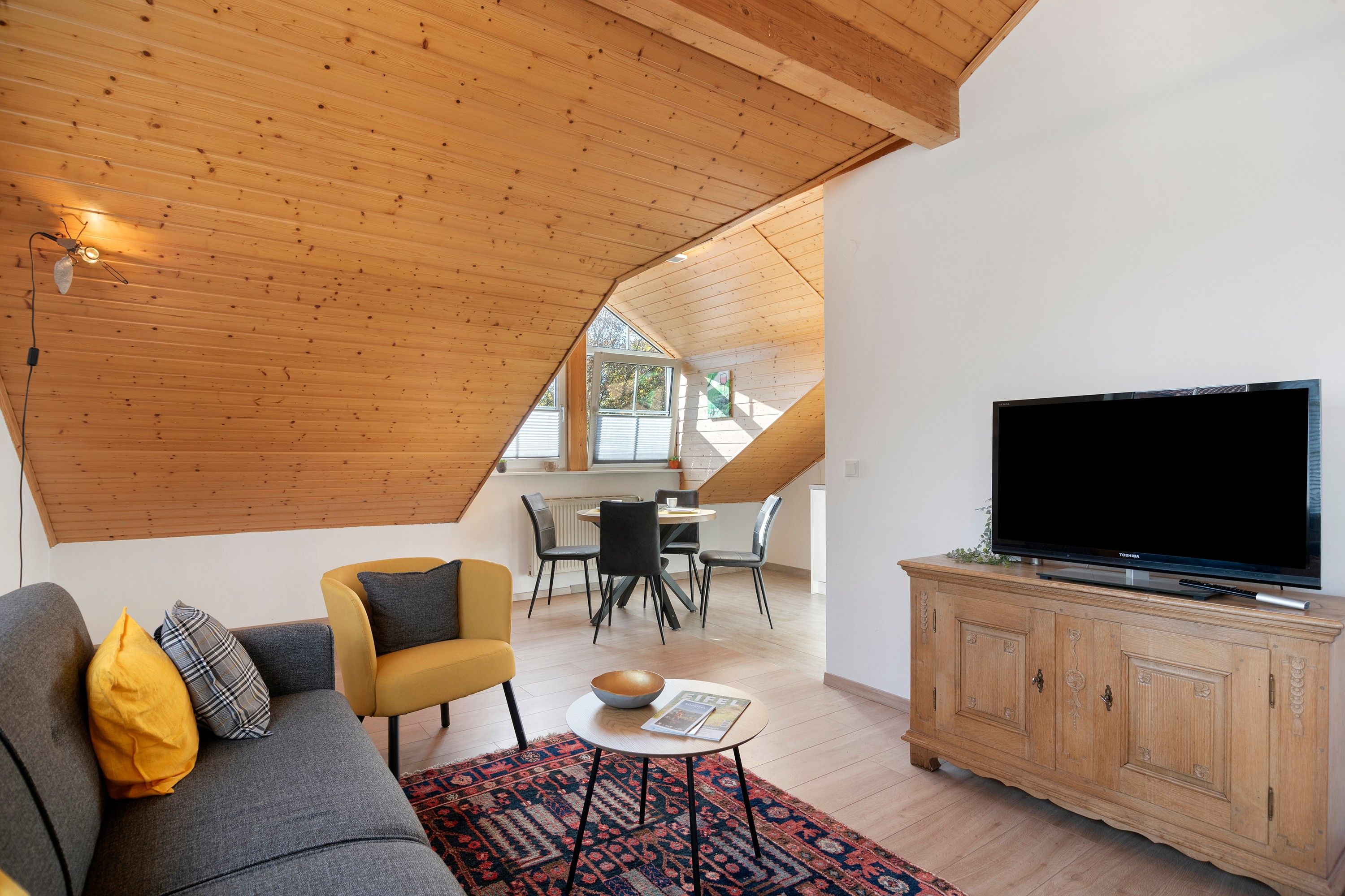 Ein modernes Wohnzimmer mit schräg versiegelter Decke, einem Fernseher auf einem Holzschrank, einer Couch mit Kissen, einem Esstisch mit Stühlen und einem gemusterten Teppich.