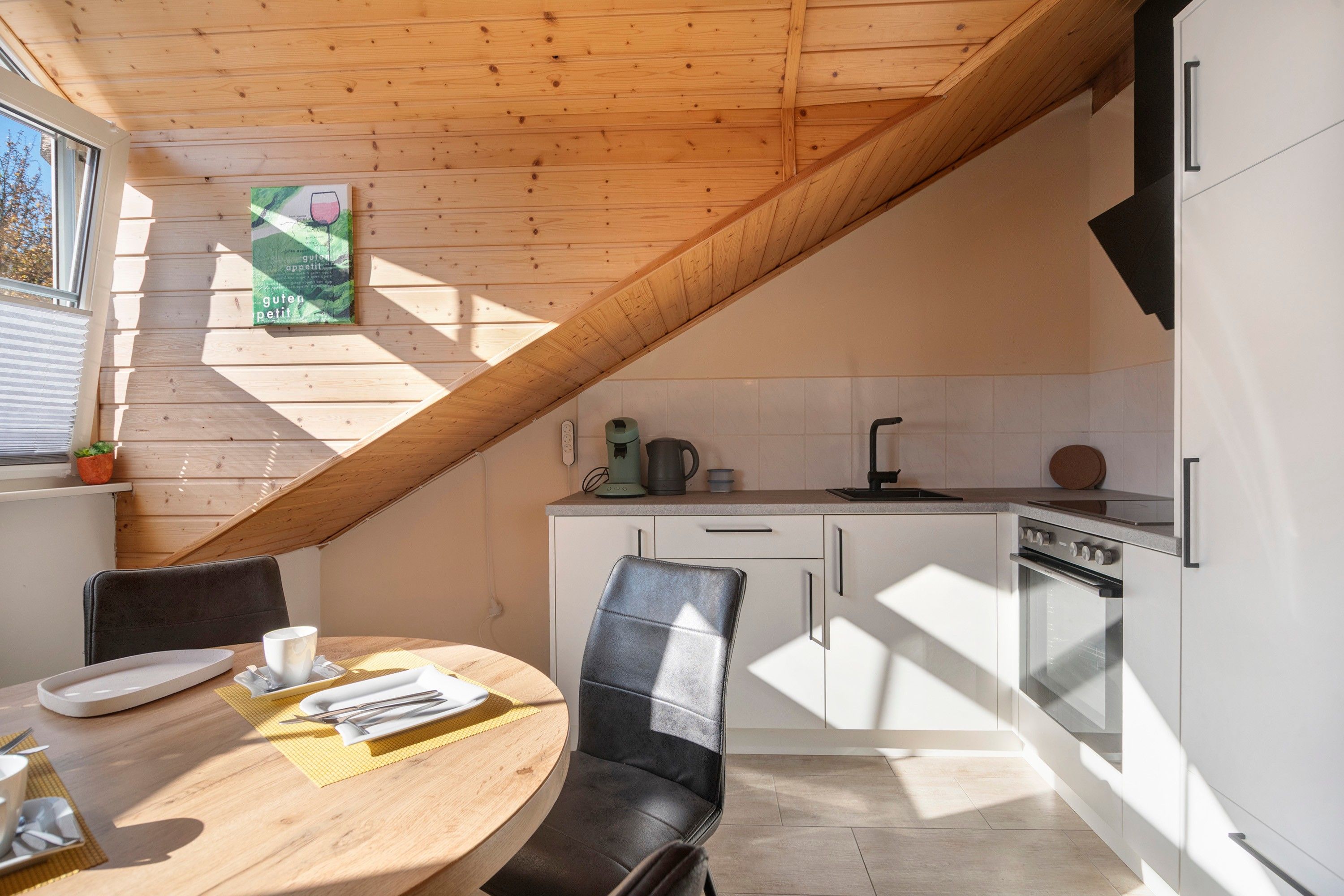 Une cuisine moderne avec un plafond en pente, un coin repas avec une table et des chaises, et une fenêtre avec des stores.