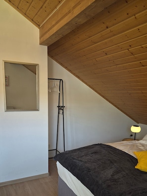 A bedroom with a sloped ceiling, a bed with a dark cover, a bedside lamp, a hanging coat rack, and a mirror on the wall.