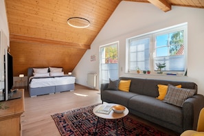 A modern loft bedroom with a sloped ceiling, a large bed, a sofa, and a small table.
